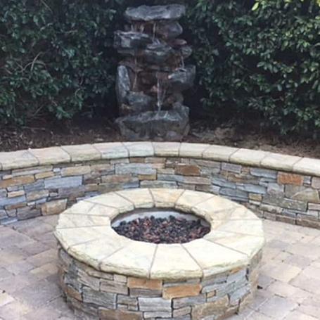A circular stone fire pit surrounded by a curved stone wall, with a decorative water feature in the background.