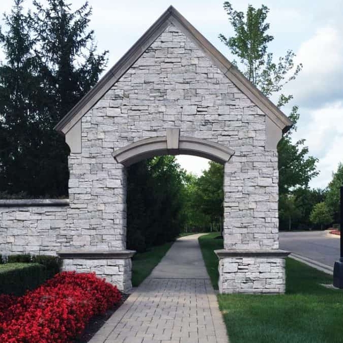 An arched stone entrance with a pathway and colorful flower beds on either side, set against a lush green landscape.