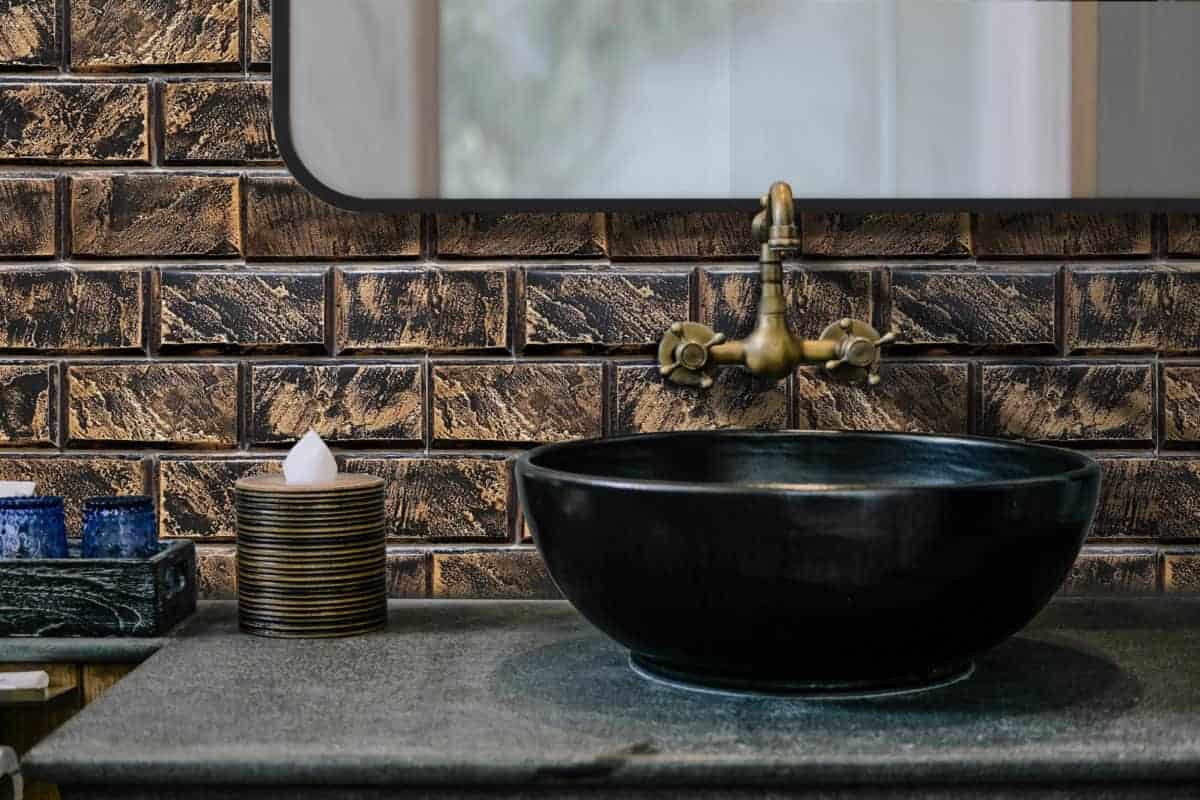 A stylish bathroom sink with a black basin, brass faucet, and a textured brick wall background. Accessories include decorative jars and a candle.