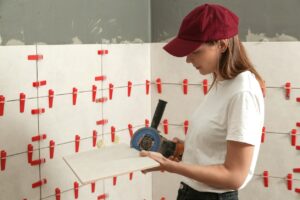 A woman in a red cap measures a tile, preparing to install it on a wall with red spacers.