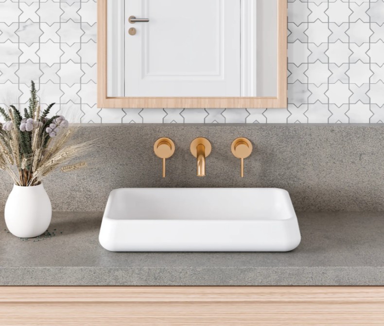 A stylish bathroom setup featuring a white rectangular sink, gold faucets, and a vase with dried flowers against a patterned tiled wall.