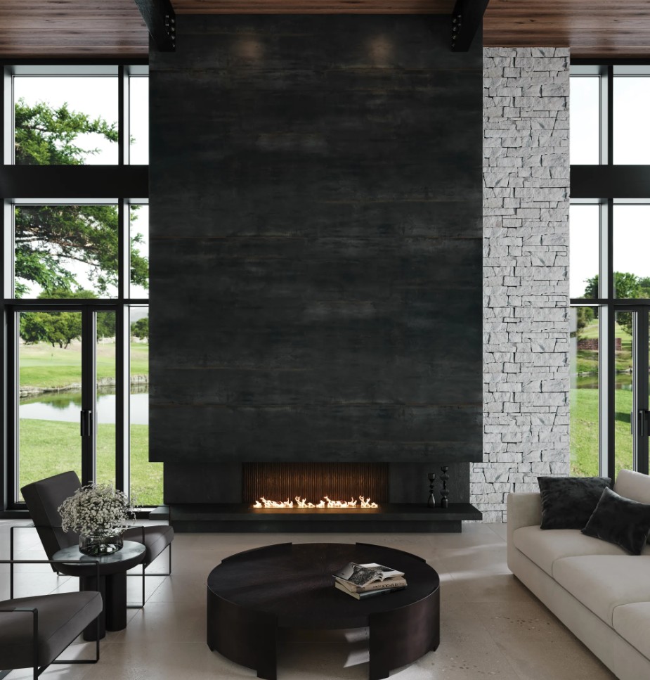Modern living room with a tall dark fireplace, white stone accents, large windows overlooking greenery, a circular coffee table, and a cozy sofa.