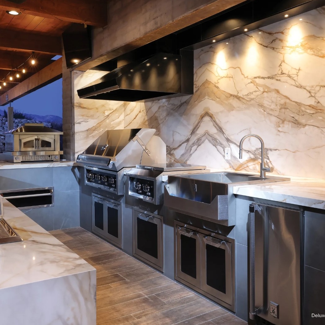 A modern outdoor kitchen features marble countertops, stainless steel appliances, and stylish lighting, set against a scenic backdrop.