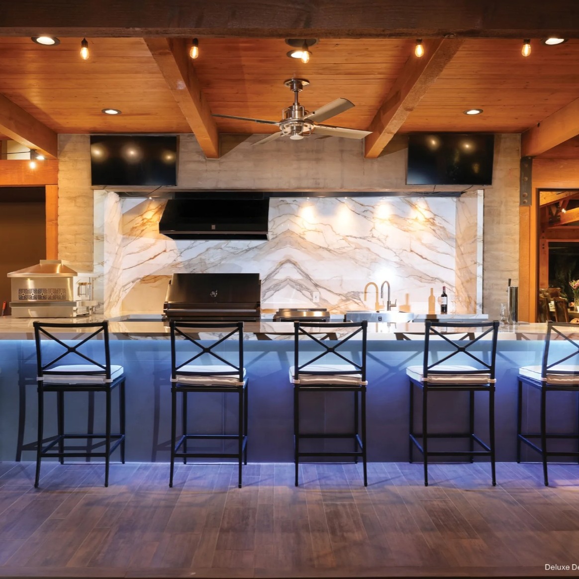 A modern kitchen bar with high stools, granite countertops, and warm wooden finishes, featuring sleek appliances and ambient lighting.