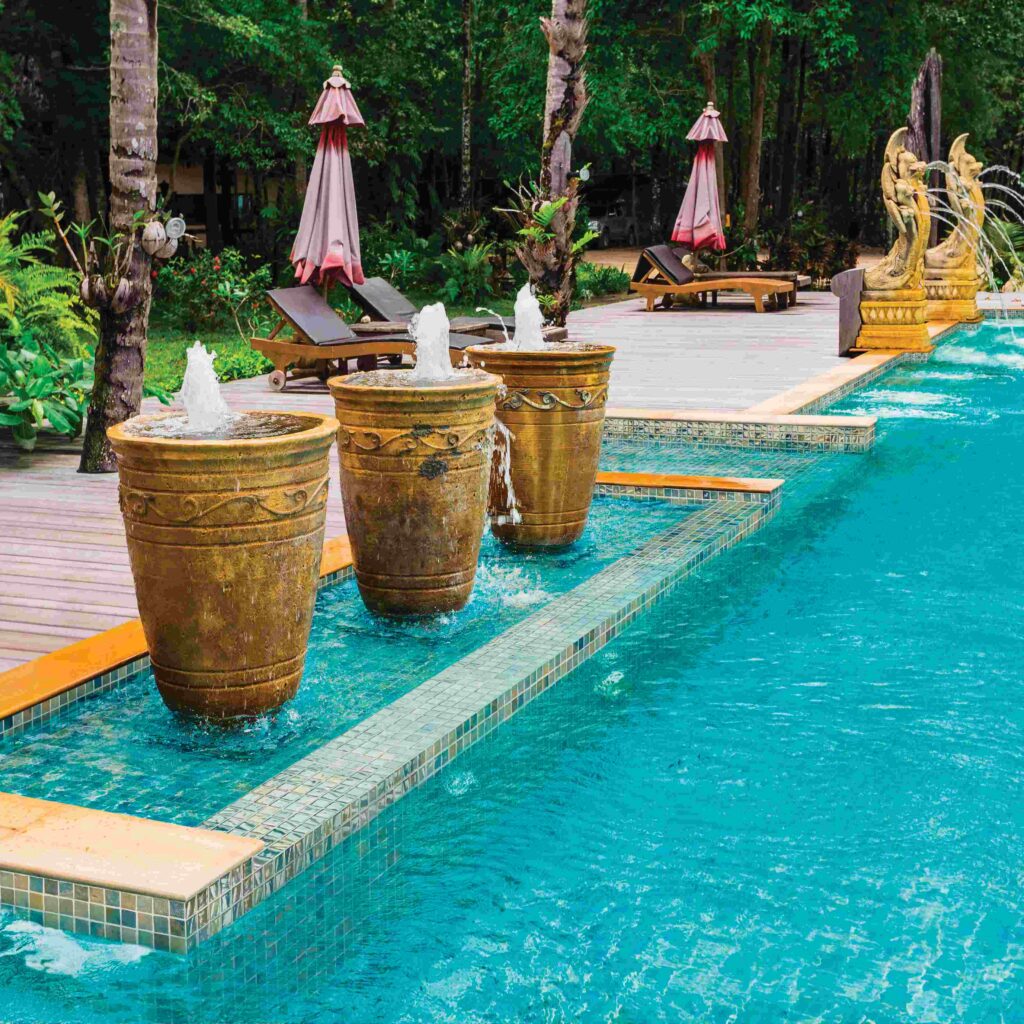 Decorative water fountains emerge from ornate golden pots alongside a tranquil blue pool, surrounded by lush greenery and relaxing chaise lounges.